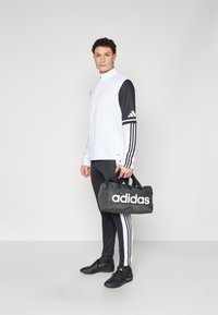Sort og hvid Adidas træningsdragt med lynlåsjakke, slim-fit bukser med tre striber og en sort duffeltaske med stort hvidt logo.