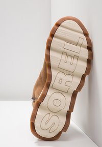 Jasnobrązowy zamszowy but z teksturowaną, brązową gumową podeszwą. Podeszwa ma ukształtowane bieżniki oraz dużym literami wytłoczoną nazwę marki "SOREL".