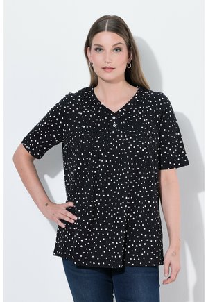 Donna con capelli castani lunghi, che indossa una camicetta nera a maniche corte con motivi a forma di cuore bianchi e jeans blu, in piedi con una mano sui fianchi.