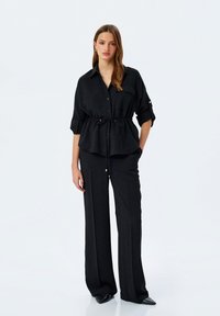 Zwarte, knoop-down overhemd met een riem in de taille, voorzien van opgerolde mouwen en een borstzak, gedragen met bijpassende wijde pantalon en puntige schoenen.