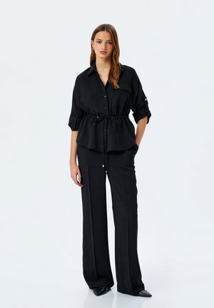 Camisa negra de botones con cintura ceñida, que presenta mangas enrolladas y un bolsillo en el pecho, combinada con pantalones de pierna ancha a juego y zapatos puntiagudos.
