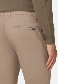 Beige casualní kalhoty s hladkým povrchem, s kapsou na zadní straně s horizontálním otvorem a černou štítkovou nálepkou.