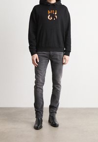 Sudadera negra con capucha y un colorido logo "HUGO", combinada con jeans grises y botas de cuero negras hasta el tobillo. Fondo liso.