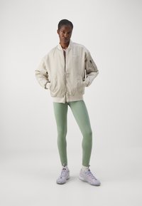 Beige bomberjakke med glidelås, sidelommer og en liten logo, kombinert med grønne tights og flerfargede joggesko.