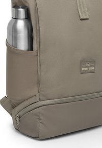 Beige hátizsák texturált anyagból, oldalsó zsebben egy ezüst rozsdamentes acél vízpalackkal, és egy kis logófolttal. Cipzár részlet alul.