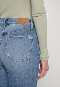 Džíny vysokého pasu modré barvy s lehkým praním, s hnědou koženou nášivkou na zadním pase a jemným prošíváním na kapsách.