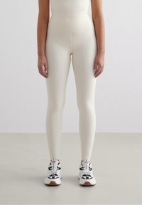 Leggings chiari in crema con una texture liscia, vita alta e una silhouette aderente, abbinati a sneaker sportive in bianco e nero.