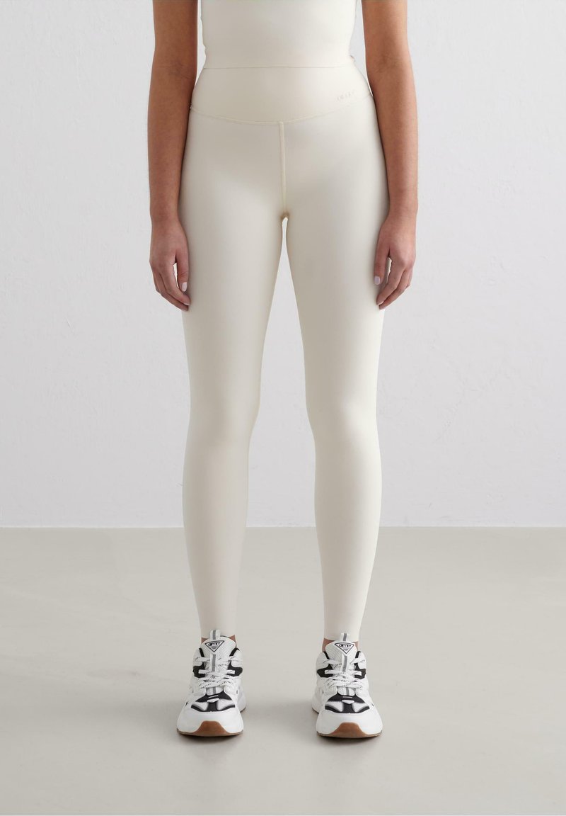 Leggings chiari in crema con una texture liscia, vita alta e una silhouette aderente, abbinati a sneaker sportive in bianco e nero.