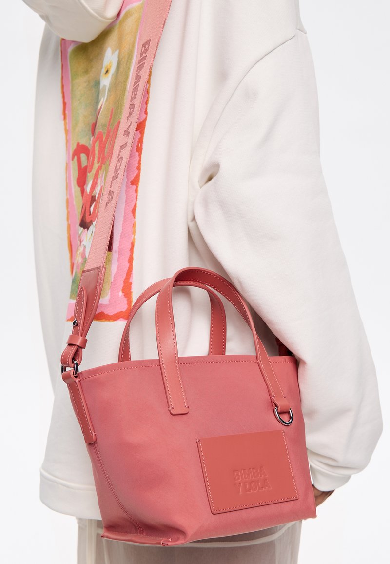 Borsa sintetica rosa con due manici superiori e una tracolla rimovibile. Presenta dettagli cuciti e una patch con logo sulla parte anteriore.
