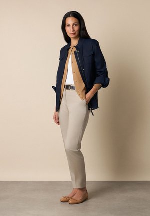 Kobieta stojąca z rękami w kieszeniach, ubrana w granatową kurtkę, beżowy kardigan, biały top, beżowe spodnie, czarny pasek i beżowe baleriny.