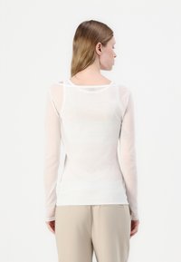 Langermet, gjennomsiktig hvit topp med avslappet passform, rund hals og myk tekstur; modell vist fra baksiden, sammen med beige bukser.