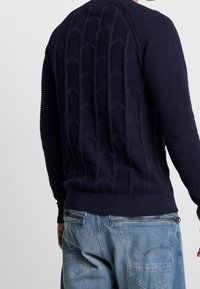 Marinblå tröja med strukturerad hexagonal design på baksidan, ribbade sektioner på ärmarna och en åtsittande midja. I kombination med ljusblå jeans.