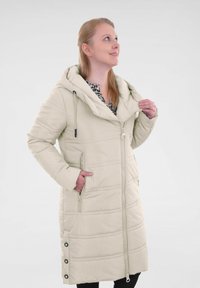 Beige pufferjack met een capuchon, ritssluiting aan de voorkant en zijzakken. Heeft een gestikt ontwerp en drukknoopaccenten aan de onderrand. Glad materiaal.