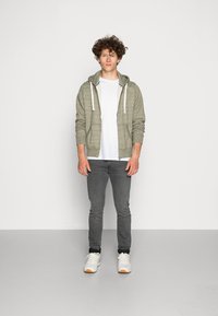 Lichtgrijze zip-up hoodie met een textuurpatroon, gedragen over een wit T-shirt, gecombineerd met donkergrijze jeans en witte sneakers met oranje accenten.