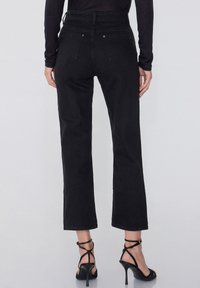 Jeans de mezclilla negra acampanados, de cintura alta con cinco bolsillos, que cuentan con una textura suave y detalles de costura en contraste. Combinados con sandalias de tacón negras.