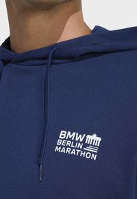 Marineblaue Kapuzenjacke mit Kordelzug, featuring ein weißes, gesticktes Logo mit der Aufschrift "BMW Berlin Marathon" und einem ikonischen Denkmal.
