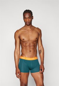 Teal boxershorts met een oranje tailleband waarop "JACK & JONES" staat. Het model heeft een atletisch postuur en kort gevlochten haar.