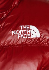Czerwona ocieplana kurtka z gładką, błyszczącą teksturą, z białym haftowanym logo "The North Face" na klatce piersiowej.