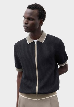 Man met kort krullend haar draagt een zwart gebreid shirt met ritssluiting met beige kraag, manchetten en tailleband, staand tegen een effen achtergrond.