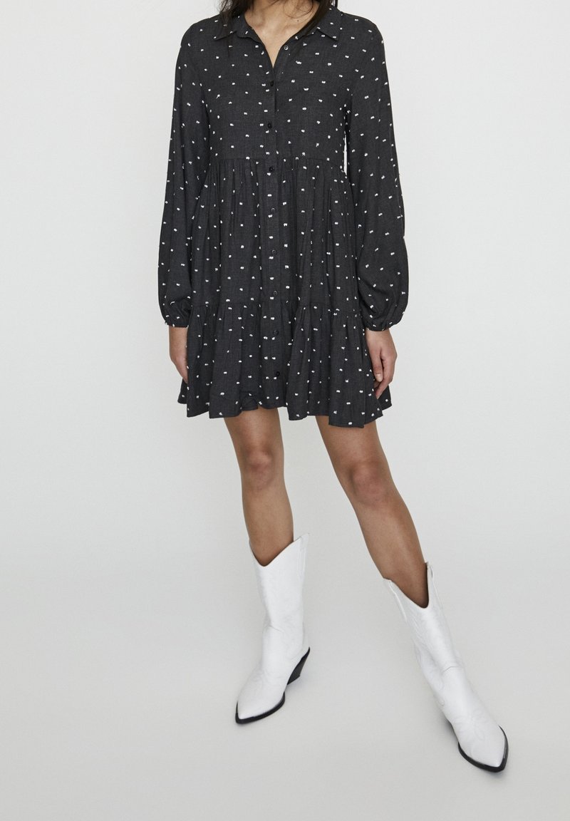 Femme portant une robe noire à pois à manches longues et des bottes blanches à bout pointu montant jusqu’au milieu du mollet, debout devant un fond uni.
