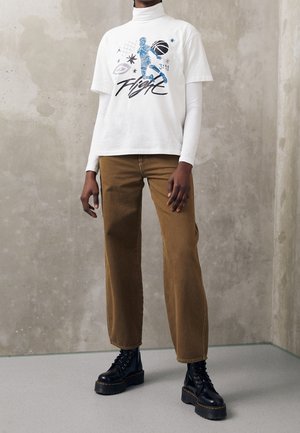 Osoba ubrana w biały golf pod białą koszulką z nadrukiem, brązowe spodnie o szerokich nogawkach oraz czarne buty na platformie na tle szarej ściany.