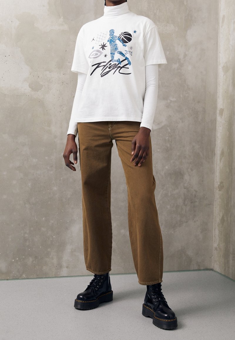 Personne portant un col roulé blanc sous un t-shirt blanc à motifs, un pantalon brun à jambes larges et des bottes à plateforme noires devant un mur gris.