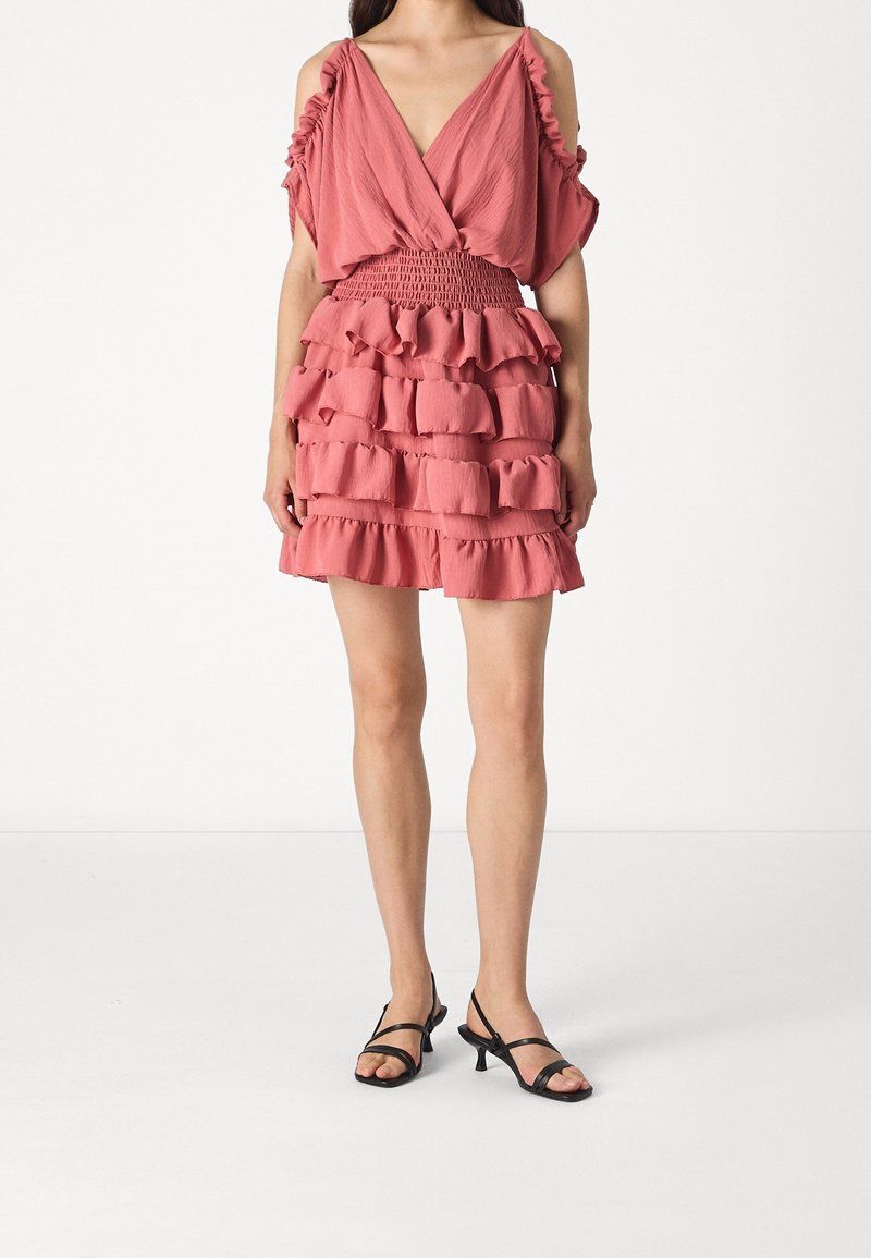 Femme portant une robe rose à volants avec des manches épaules dénudées et une taille smockée, associée à des sandales noires à talons bas et brides.