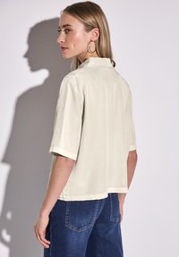 Camicia beige chiaro a maniche corte con colletto, caratterizzata da un taglio dritto e spacco laterale. Indossata con jeans blu scuro, mostrando una vista da dietro.