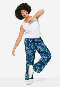 Top bianco senza maniche con dettagli di bottoni, abbinato a pantaloni blu con motivo paisley verde; indossato con calzature bianche.