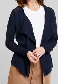 Granatowy kardigan z otwartym przodem, z siateczkowymi detalami na rękawach i miękką, ribbed teksturą. Noszony na białej bluzce.