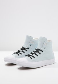 Jasnoniebieskie sneakersy typu high-top z czarnymi sznurówkami, białą gumową osłoną palców oraz teksturowanym materiałem z płótna. Czarny akcent logo na pięcie.