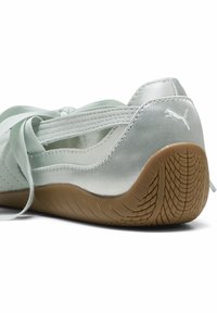 Ασημένιο μπαλαρίνα Puma με σατεν κορδόνια και υφασμένο καφέ καουτσούκ πάτο, ορατό από πίσω και πλάγια γωνία.