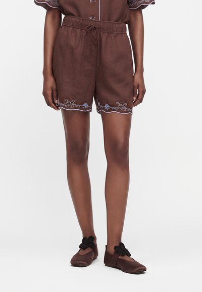 Short marron avec une taille élastique et une broderie florale à l’ourlet, porté avec des chaussures marron assorties avec des nœuds noirs.