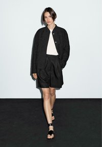 Zwarte gewatteerde jas over een wit shirt, gecombineerd met zwarte shorts tot de knie. Open-teense zwarte sandalen maken de outfit compleet.