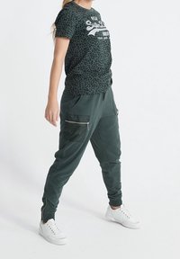 Zielona koszulka w panterkę z białym logo, w połączeniu z ciemnozielonymi dresami z zamkami błyskawicznymi i białymi sneakersami.