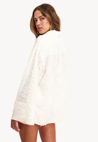 Chaqueta blanca texturizada con un corte holgado, que presenta un diseño con flecos, mangas largas y un dobladillo recto. La tela parece suave y ligera.