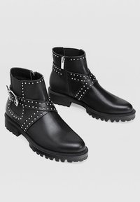 Bottes en cuir noir à la cheville avec des clous en argent et un design à sangle croisée. Comprend une fermeture éclair sur le côté et une semelle robuste pour une meilleure adhérence.