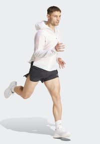 Witte, lichte hardloopjack met een capuchon en een rits aan de voorkant, gecombineerd met zwarte shorts en lichtgrijze sport schoenen, in een hardloophouding.