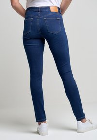 Jeans de mezclilla azul ajustados con bolsillos traseros, una etiqueta de cuero marrón en la cintura y una textura suave. Llevados con zapatillas blancas.