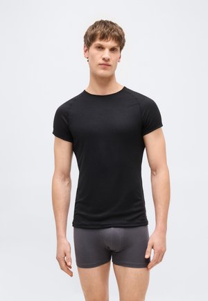 Młody mężczyzna stojący przodem, ubrany w dopasowaną czarną koszulkę z krótkim rękawem i ciemnoszare bokserki na jednolitym tle.