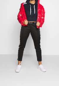 Giacca puffer rossa sopra una felpa cropped blu marina, abbinata a joggers neri a gamba affusolata con strisce bianche ai lati e sneakers bianche.