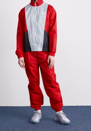 Personne portant une veste coupe-vent Nike rouge, noire et grise avec un pantalon rouge assorti et des chaussures de sport grises, debout sur un tapis bleu.