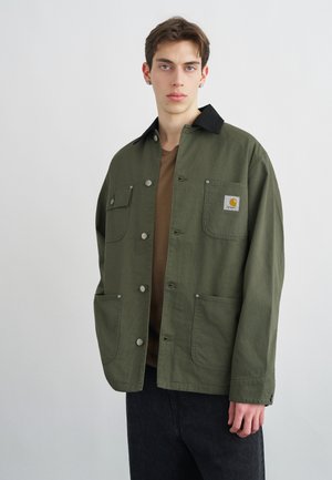 Νέος άνδρας με κοντά καστανά μαλλιά φοράει μπουφάν χρώματος ελιάς με μαύρο γιακά και μπροστινές τσέπες, στέκεται μπροστά σε απλό ανοιχτόχρωμο τοίχο.