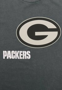 Tmavě šedé tričko s velkým černobílým oválným logem, které obsahuje písmeno "G" a slovo "PACKERS" v tučném bílém písmu. Bavlněný materiál, hladká textura.