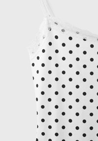 Camisole blanche à pois noirs avec bordure en dentelle le long du décolleté et fines bretelles sur fond uni.