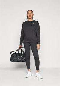 Felpa nera con logo, leggings neri, sneaker bianche e borsa da viaggio nera. Tessuto liscio, design aderente e accenti bianchi a contrasto.