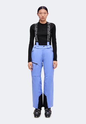 Femme debout portant un haut noir à manches longues, un pantalon de ski bleu clair avec des bretelles de marque, et des chaussures de ski noires, faisant face à l'avant sur fond clair.