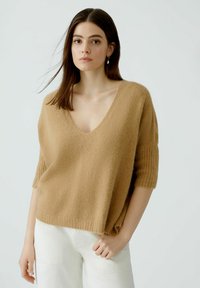 Hellbrauner, kurzärmliger Pullover aus weichem, fuzzy Material mit tiefem V-Ausschnitt und gerippten Akzenten, kombiniert mit weißen Hosen.