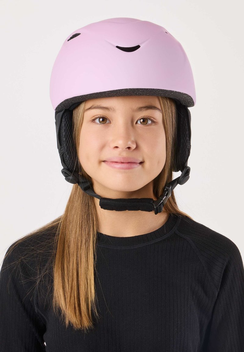 Pink helmet with a smiling face design, matte finish, black adjustable chin strap, worn by a young person in a black ribbed top.
