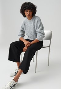 Grijze fuzzy trui met geribbelde halslijn, zwarte wijde broek en witte sneakers met zwarte accenten, zittend op een chroom stoel.